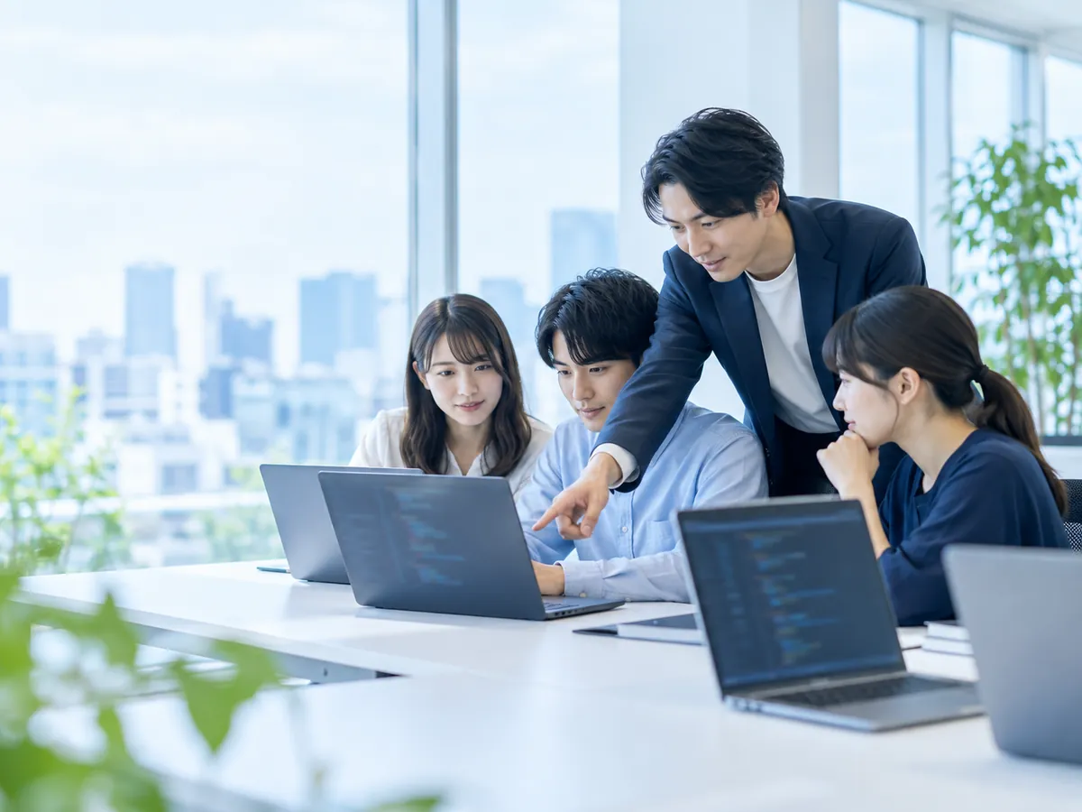 会議室でノートパソコンに向かう新入社員に、先輩社員が画面を指さして教えている様子